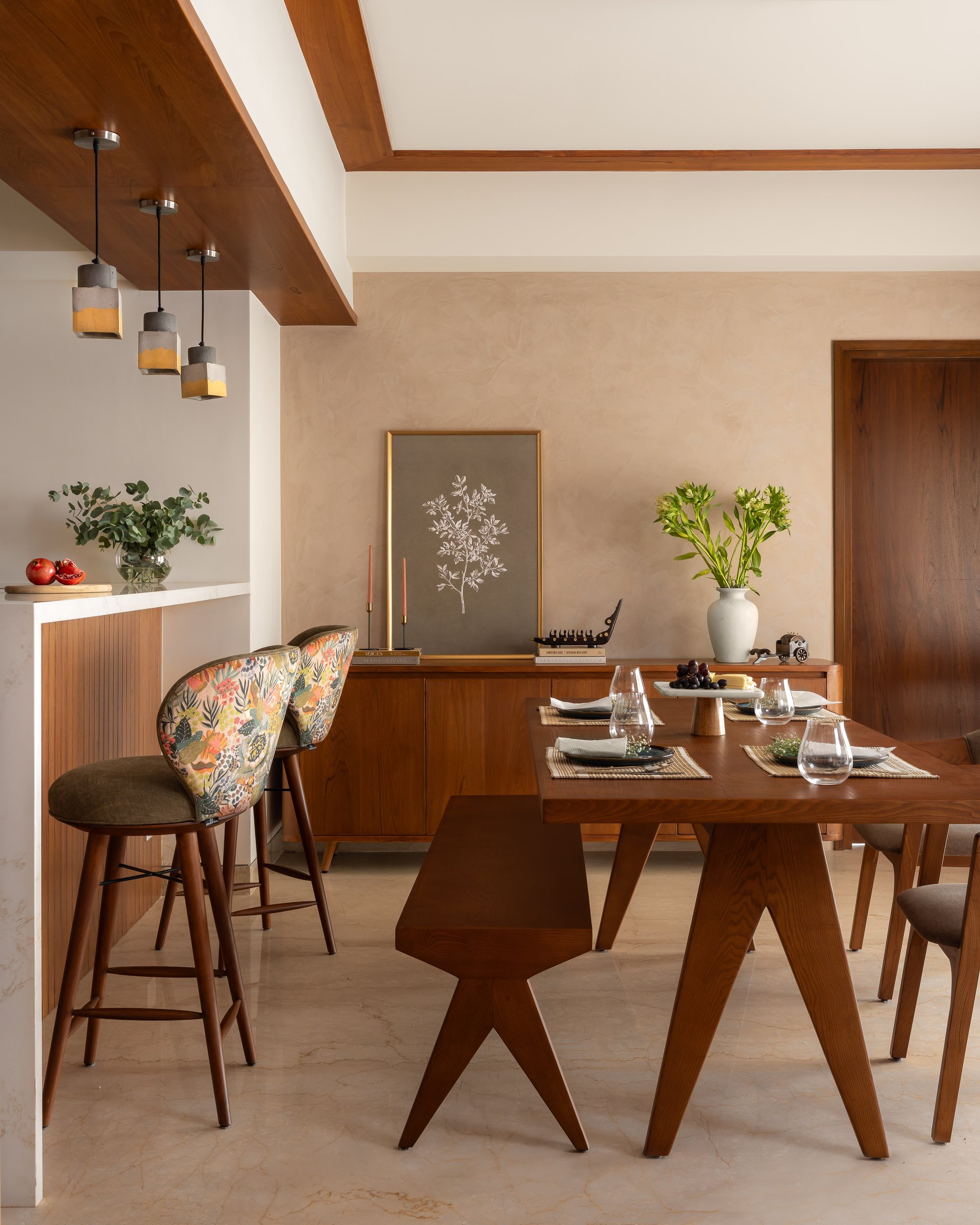 The dining area, with its Chandigarh-referencing timber dining chairs and cluster of concrete pendants anchoring the transition between kitchen counter and table