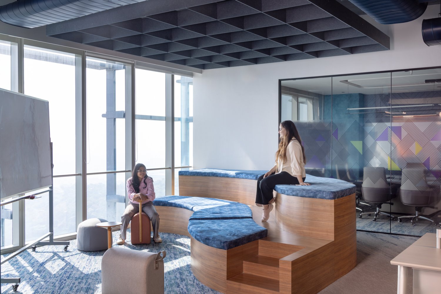 The collaboration pocket: a tiered seating platform in pale wood and mottled blue cushioning curves through the room beneath a charcoal acoustic ceiling grid