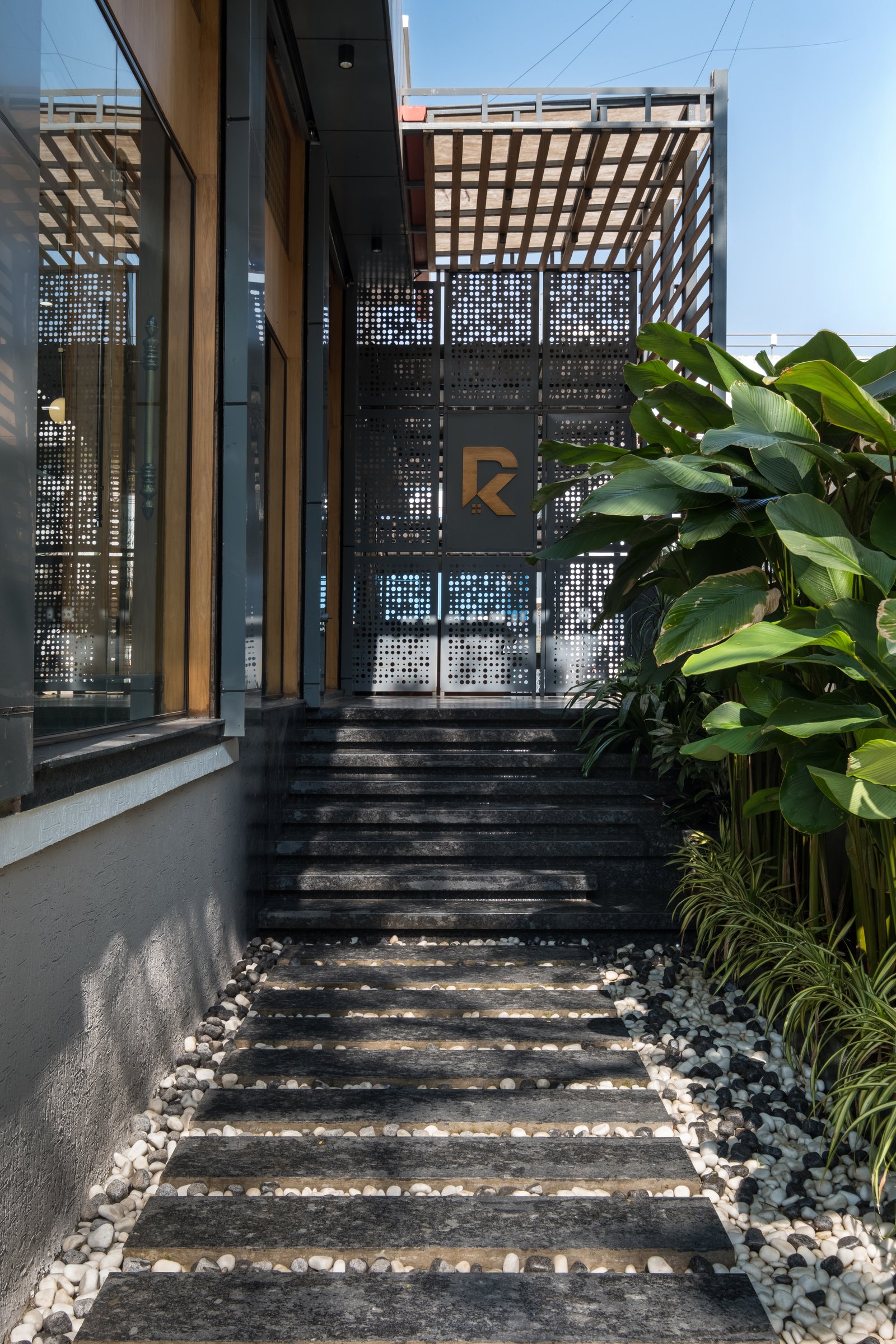 The entry sequence: stone treads in white pebble beds, with a pergola and perforated metal screen filtering light before the door is reached