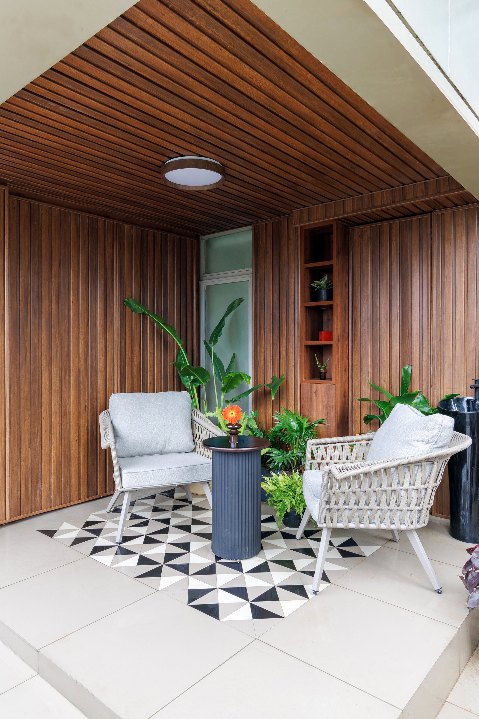 The balcony, lined in warm teak boarding, reads as an entirely different climate; a graphic tile carpet anchors a pair of rope-weave chairs around a drum table