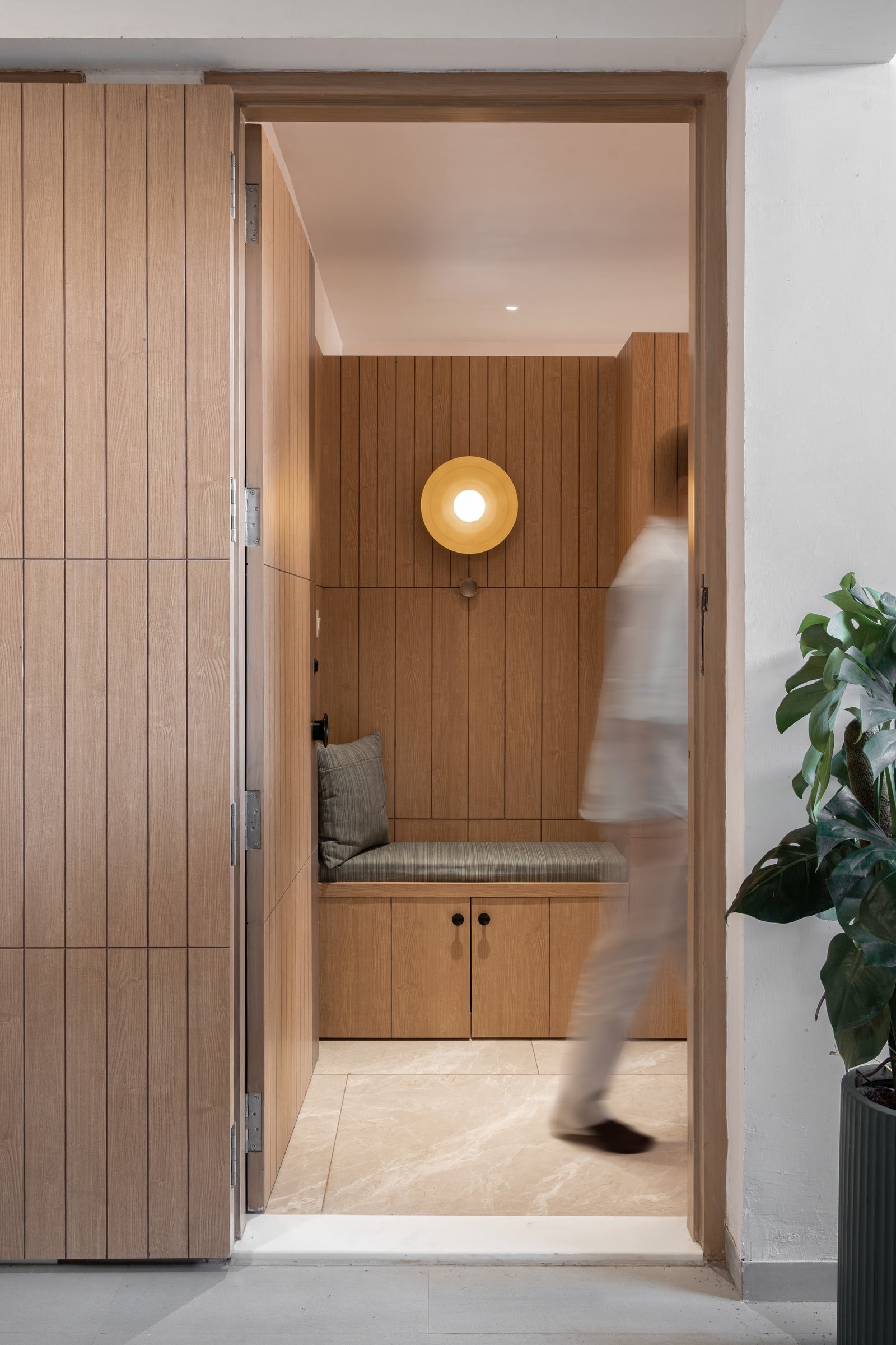 The foyer, lined in vertical wood boards with a built-in bench and circular sconce, reading as the apartment in miniature