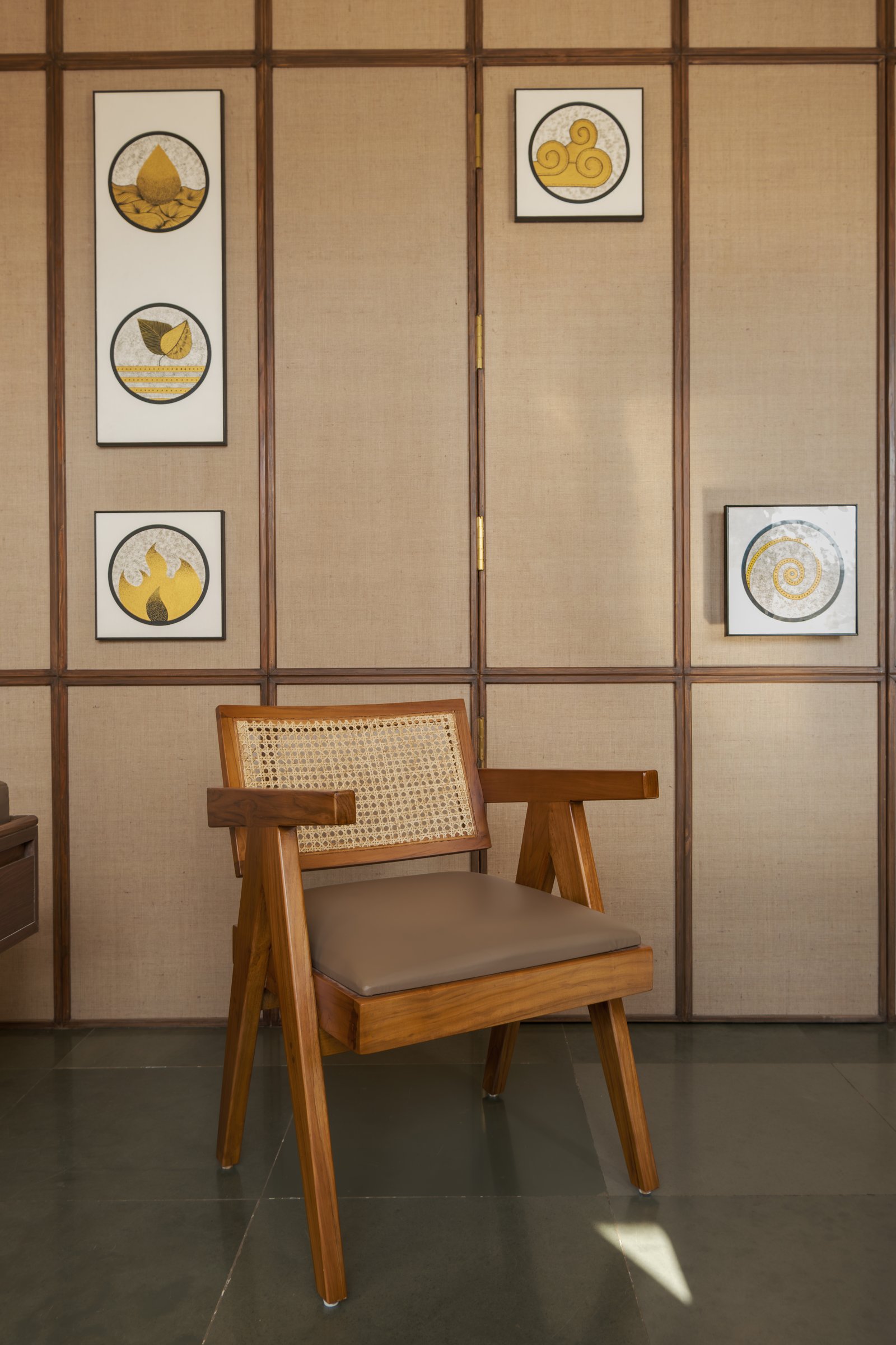 A cane-and-teak armchair against the jute panelling, the project's material vocabulary in miniature