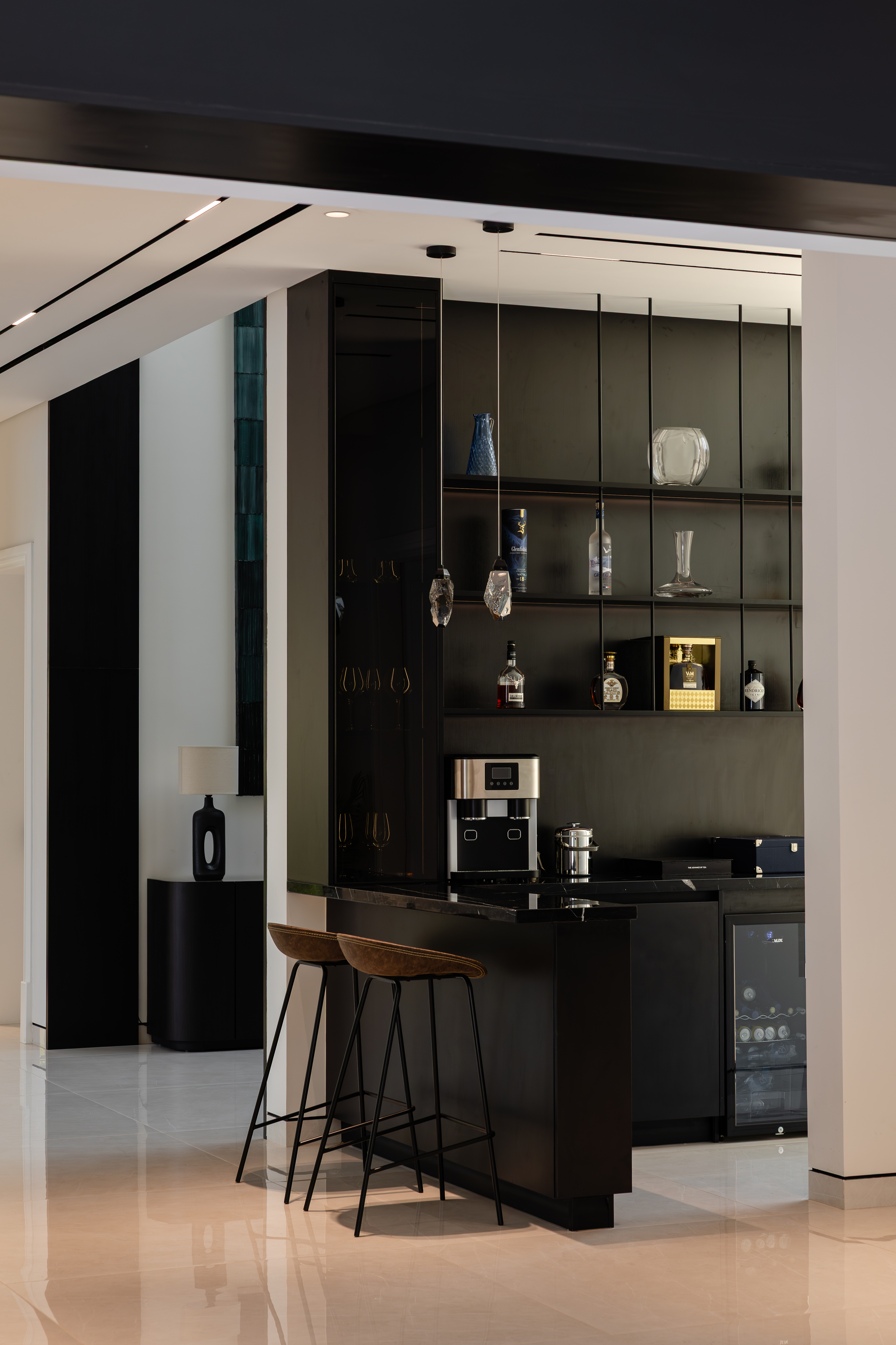 Dark built-in home bar with glass shelving, stools, and stone countertop off a hallway