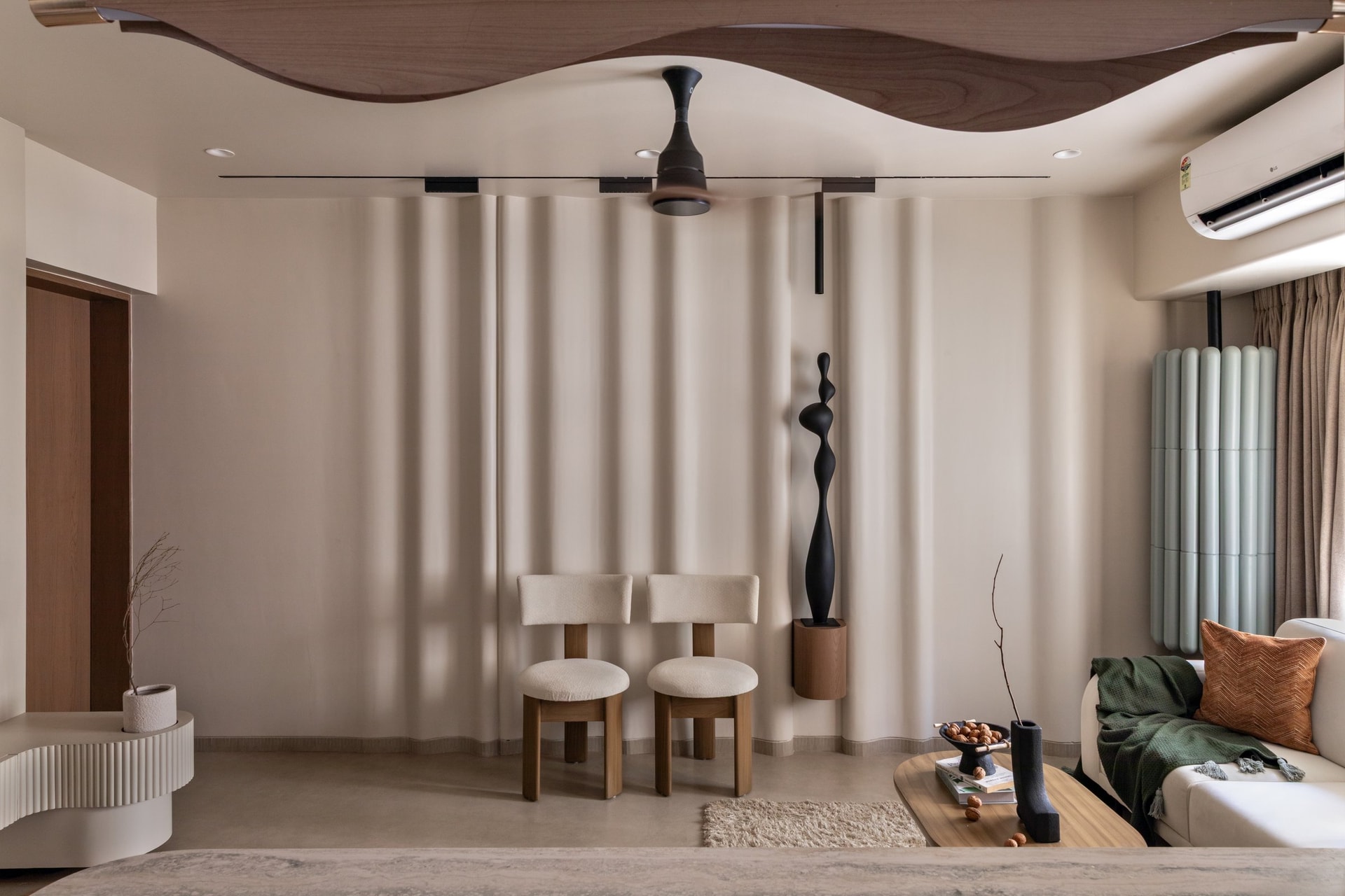 A sculpted wooden ceiling element hovers above two bouclé chairs and a slim wall-mounted plinth holding a dark figure