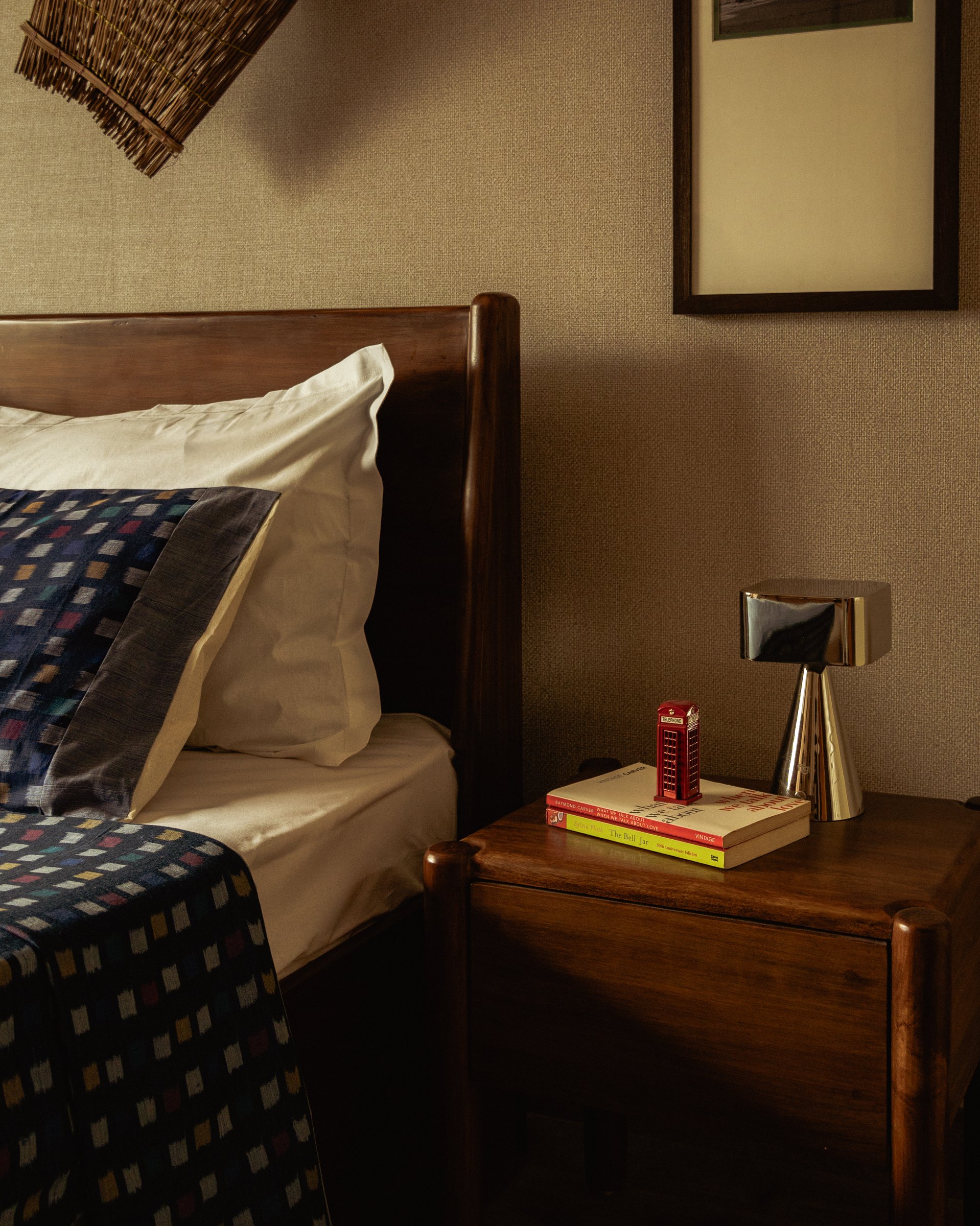 The nightstand, where Assamese heritage above the bed meets international paperbacks and a small red telephone-box figurine at the bedside