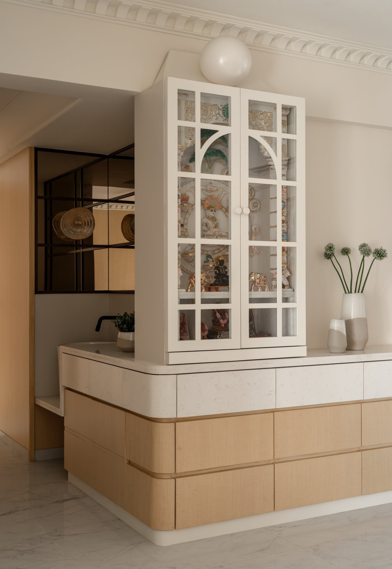The puja cabinet, its ivory arched glazed doors framing carved figures above a stepped blond wood base