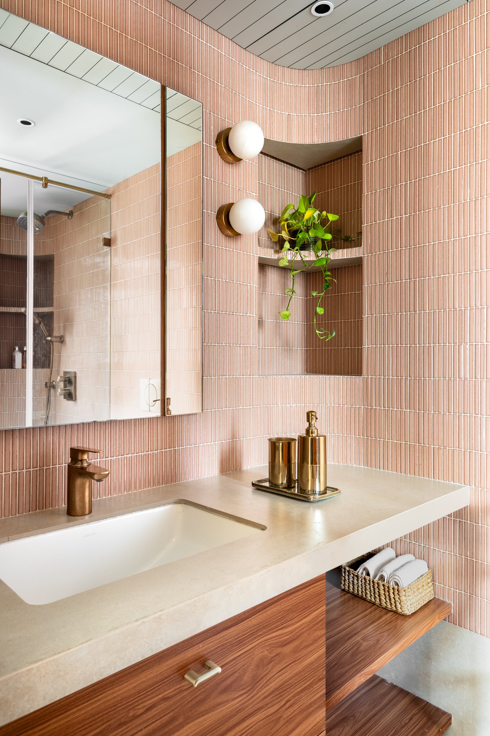 A second bathroom wrapped in stacked pink tiles that curve into the ceiling, with a concrete-toned vanity above walnut cabinetry