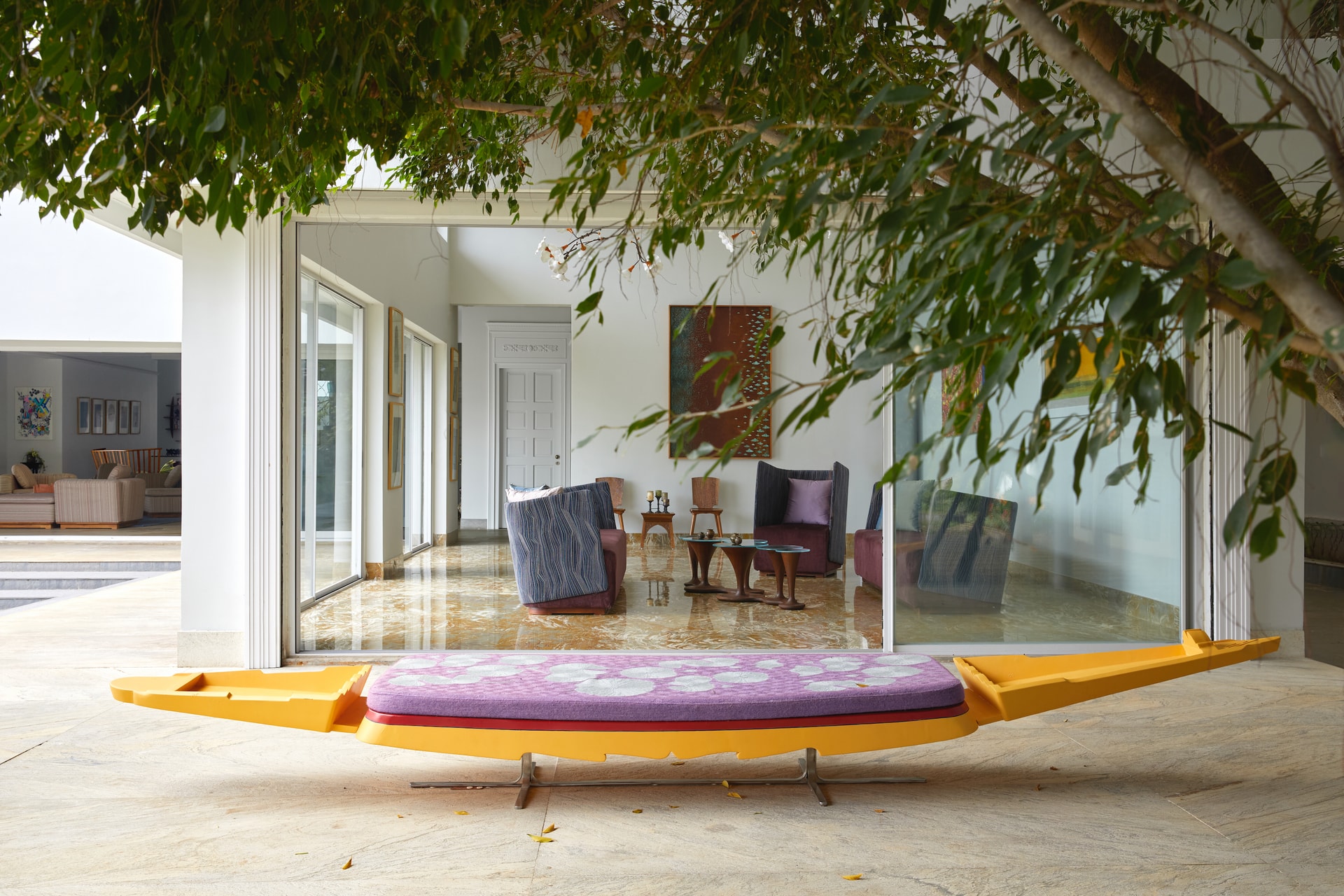 Yellow boat-shaped daybed with floral cushion under a tree outside a glass-fronted living room