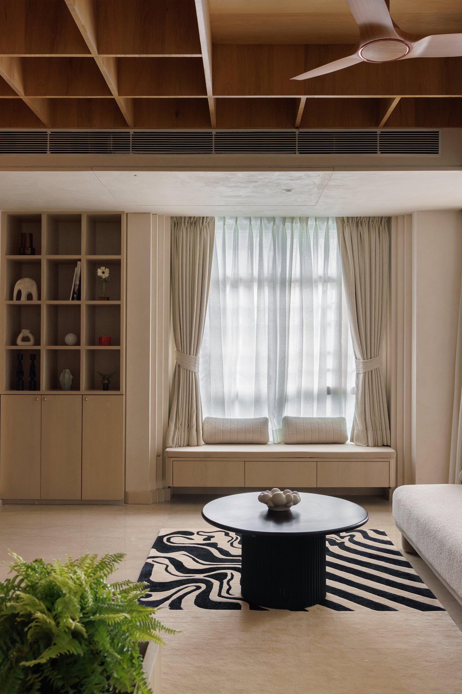 The window edge, where a built-in bench tucks beneath sheer drapery and a tall display unit echoes the coffered ceiling above