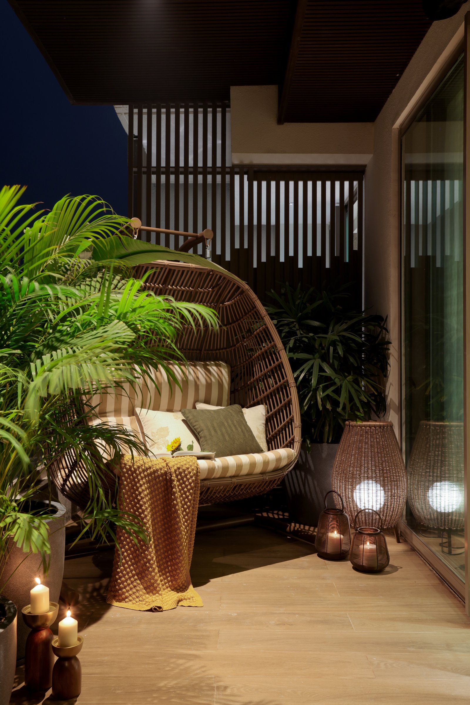The balcony at dusk: a rattan hanging chair, lanterns, and clustered palms close the tour on an informal note