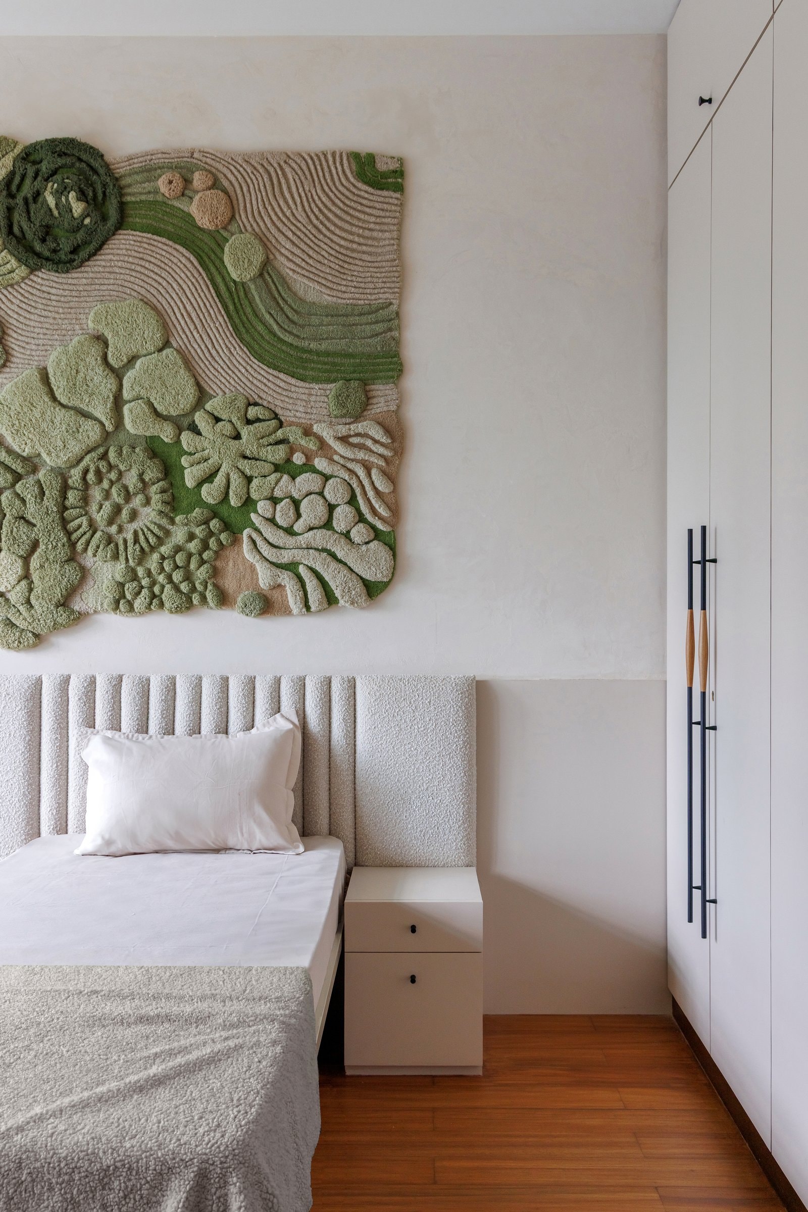 In a child's bedroom, a tufted textile artwork in mossy greens hangs above an upholstered headboard, the plaster wall behind kept deliberately quiet