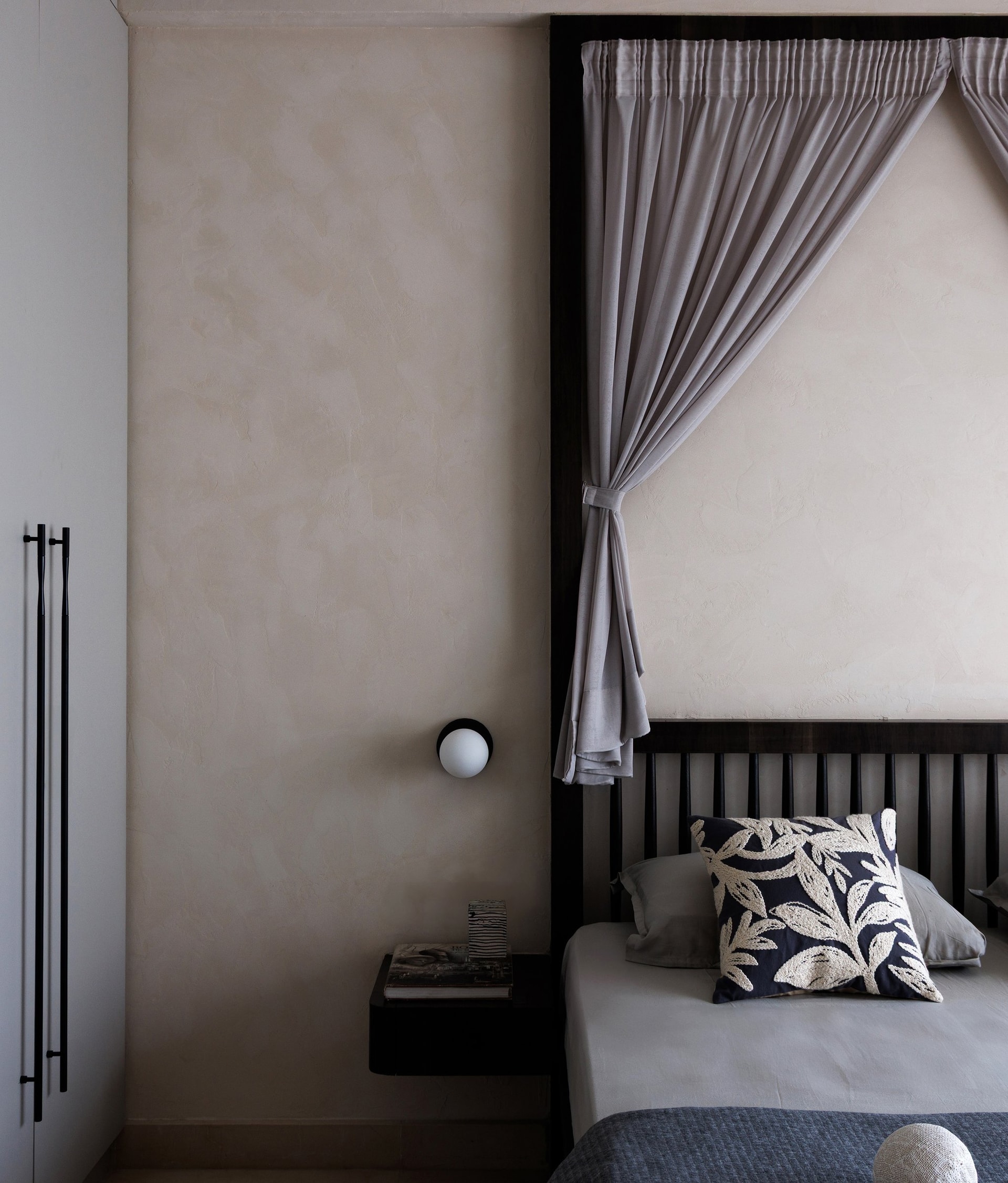 The guest room takes a graphic tone: a dark spindle headboard meets a textured plaster wall and a slim wall-mounted reading light