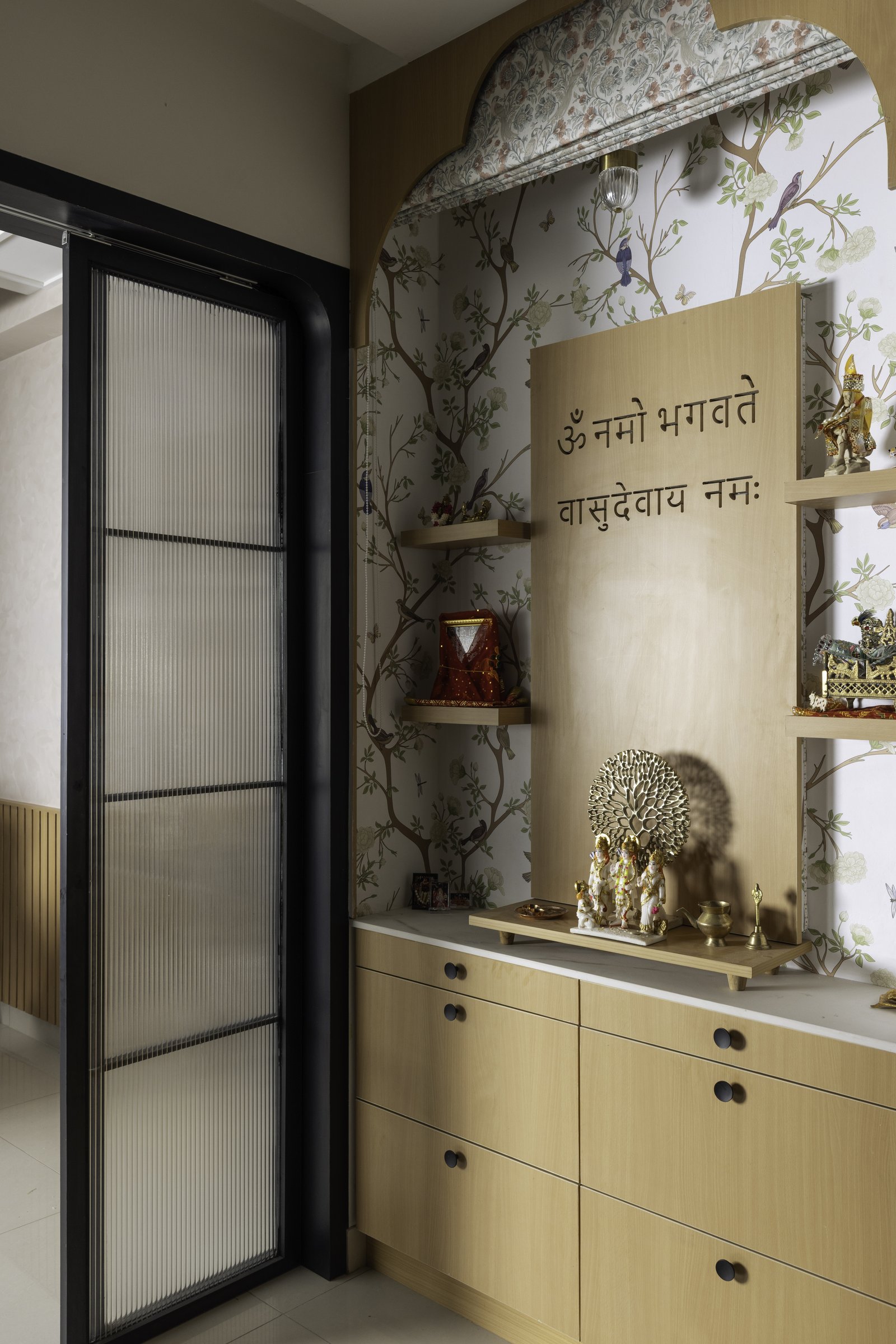 The puja room behind a fluted glass sliding door, its chinoiserie wallpaper and Sanskrit invocation marking the home's quietest pause