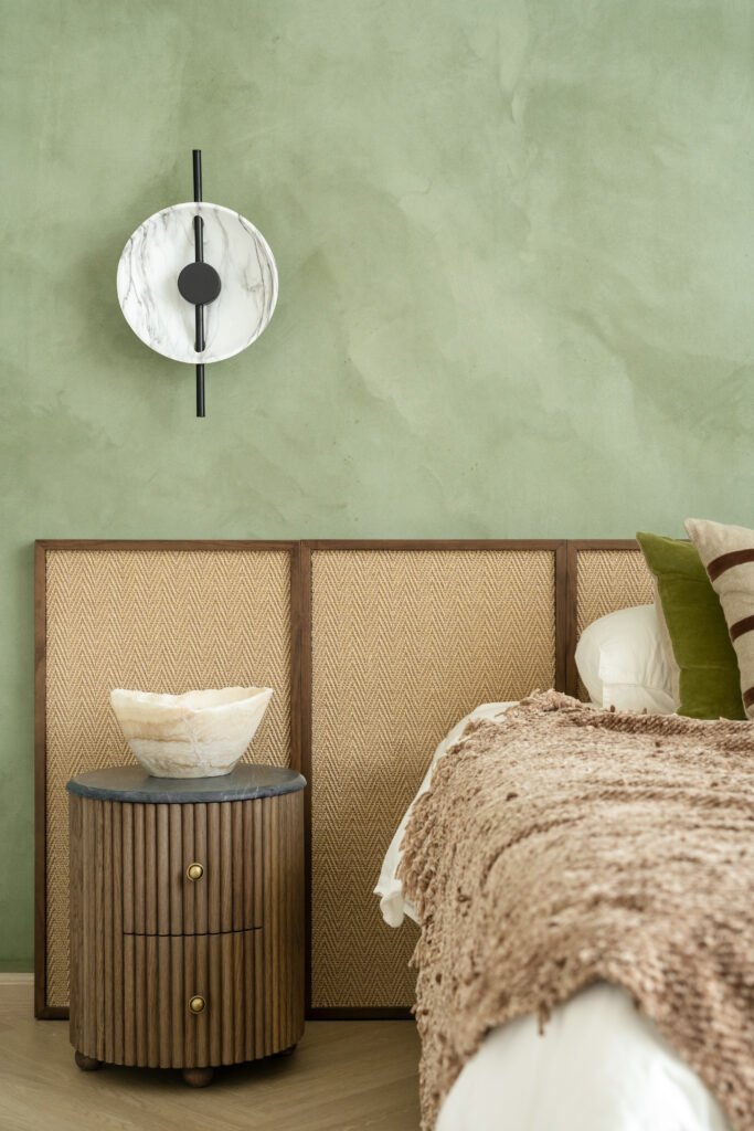 A second bedroom in mineral-green plaster, with a herringbone cane headboard and a marbled-disc sconce as its single piece of jewellery