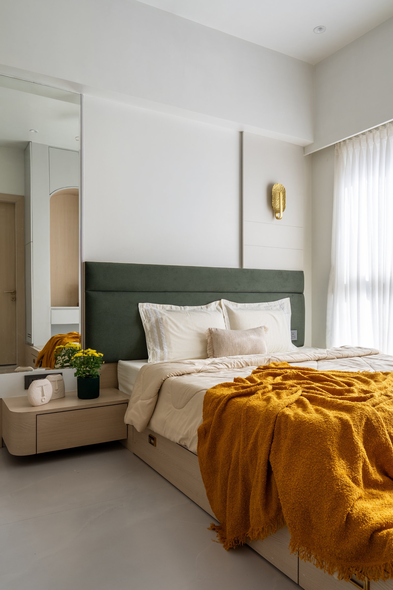 The master bedroom, anchored by a deep sage-green upholstered headboard, a brass sconce, and a mirrored panel that bounces light through the plan