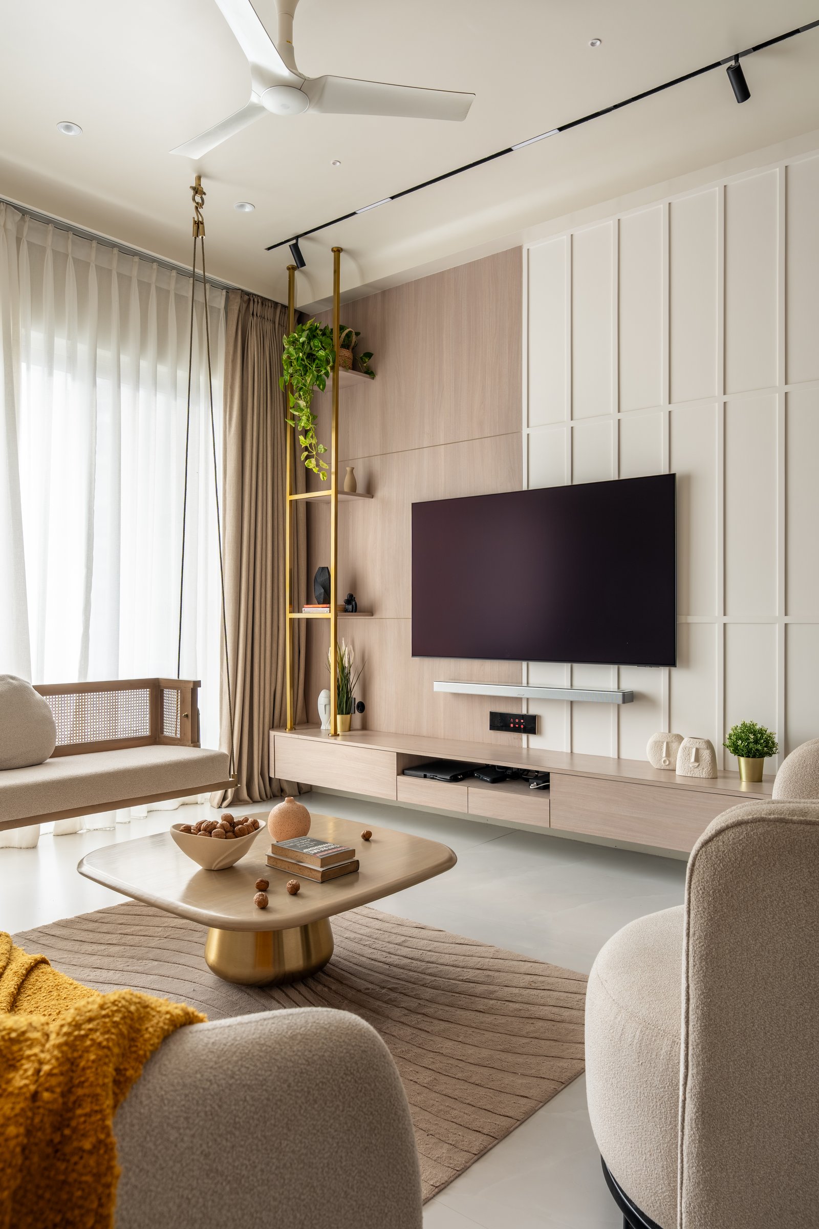 The media wall, where warm wood panelling and cream board-and-batten meet, softened by a brass shelving column threaded with greenery