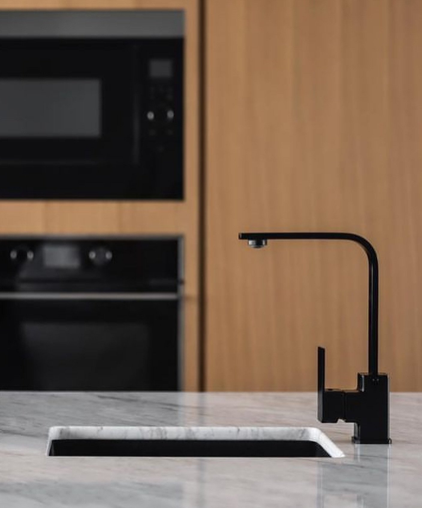A square-profile black faucet against pale veined stone, with recessed black appliances softened into the background.