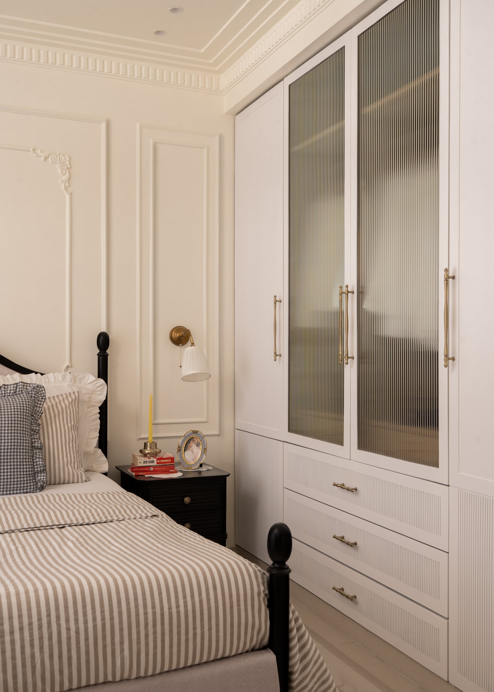 A tall wardrobe with fluted glass doors and a dark bedside chest complete the bedroom's composition